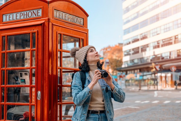Mujer turista con cámara fotográfica junto a una cabina de teléfono londinense mirando los edificios hacia arriba.