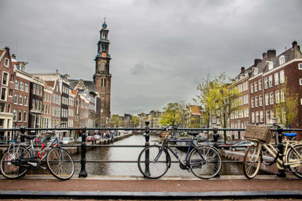 Vista de un canal de Ámsterdam con bicicletas apoyadas sobre un puente y la iglesia Westerkerk al fondo en un día nublado.