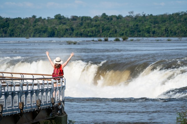 Mujer turista feliz alzando los brazos desde una pasarela frente a la Garganta del Diablo, Cataratas del Iguazú, Argentina.