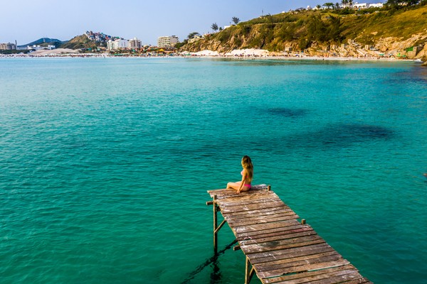 Vista aérea de Praia Grande y Deck dos Pescadores en Arraial do Cabo, Río de Janeiro, con un hermoso mar con diferentes tonos azules y una mujer en bikini sentada en el muelle.