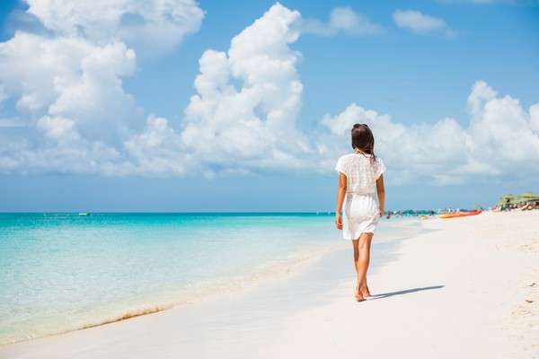 Mujer con vestido blanco caminando descalza por una playa caribeña de arena blanca.