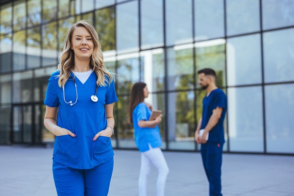 Profesional de la salud en las afueras de un centro médico con otros profesionales de la salud charlando de fondo.