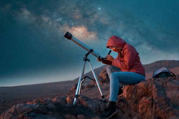 Mujer abrigada sentada en una roca usando un telescopio en el desierto por la noche.