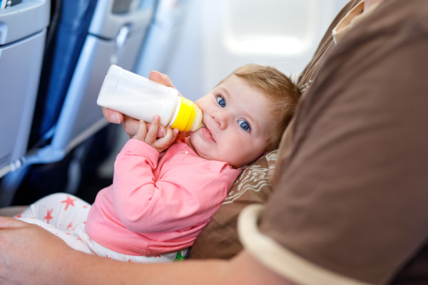 Bebé con biberón en la boca mirando a cámara en el regazo de un familiar en el asiento de un avión.