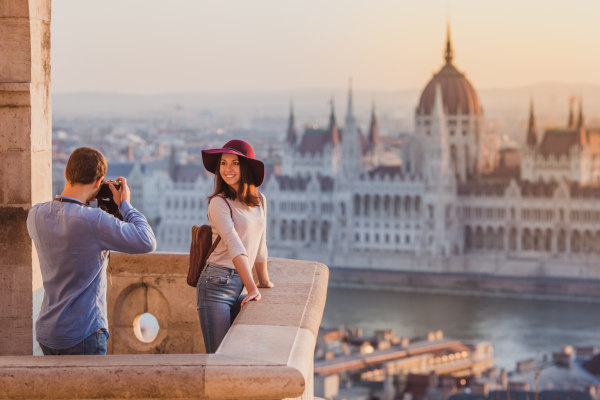 Hombre tomando fotografía de una mujer turista en el Bastión de los Pescadores con el edificio del Parlamento de Hungría de fondo.