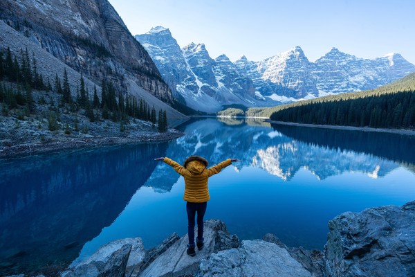 Mujer turista con abrigo alzando los brazos sobre una roca en las orillas del Lago Moraine, Canadá.