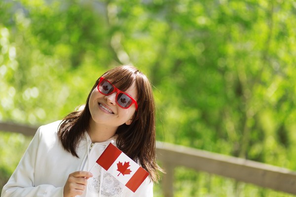 Niña con gafas de sol rojas y camisa blanca sonriendo y sosteniendo una pequeña bandera de Canadá sobre un fondo con vegetación borroso.