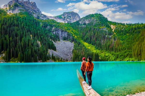 Jóvenes turistas parados sobre un tronco que se hunde en el lago Joffre, Canadá, admirando el paisaje montañoso de fondo.