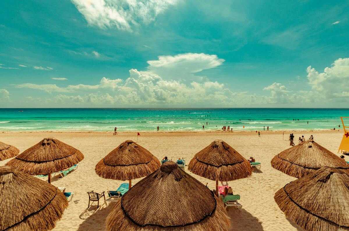 Imagem de praia em Cancún com areia clara, mar turquesa e guarda-sóis de palha, em artigo de blog da Assist Card sobre a melhor época para ir para Cancún