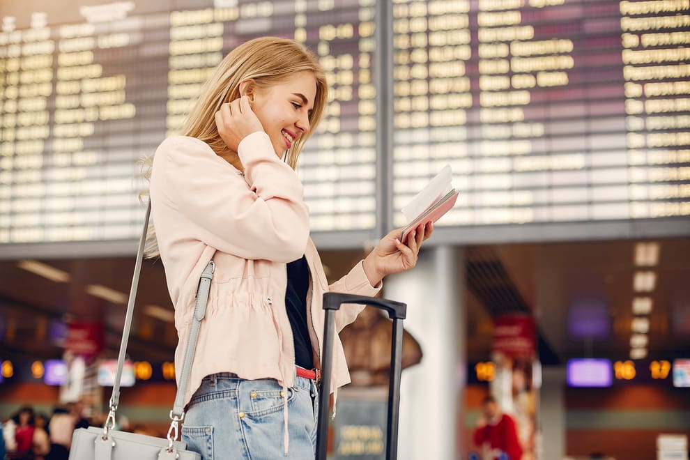 Imagem de mulher em pé com uma mala, em um aeroporto, olhando para o celular, em artigo de blog que fala sobre checklist de viagem

