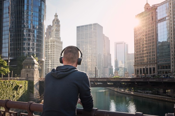 Hombre de espaldas con auriculares puestos apoyado en una barandilla de un puente sobre el río Chicago y paisaje urbano de fondo durante un día soleado.