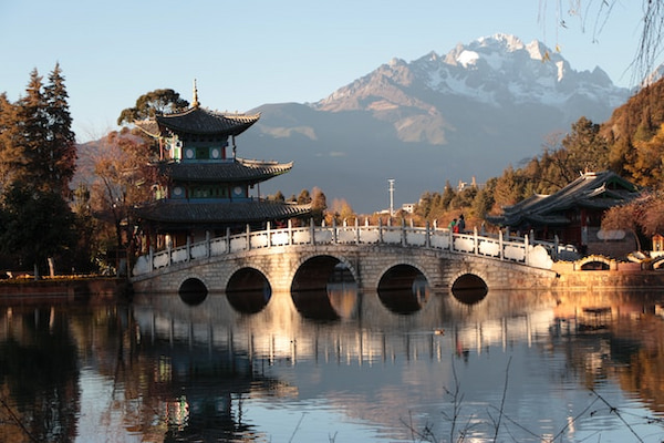 Templo e ponte de concreto cinza durante o dia, com montanha ao fundo, em Lijiang, na China.