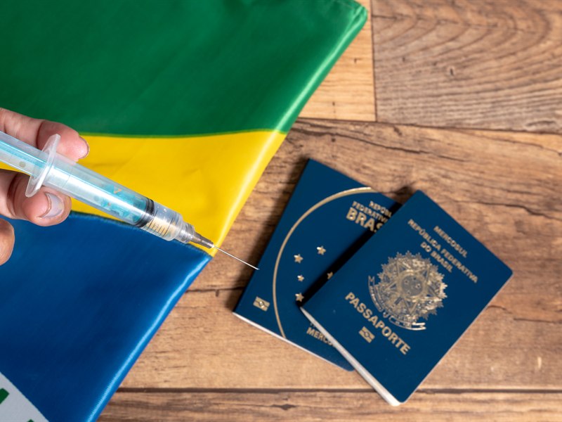 Passaportes ao lado de uma seringa de vacina e da bandeira do Brasil, representando os documentos necessários para viajar ao exterior com o certificado internacional de vacinação.