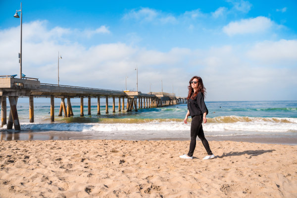 Mujer con ropa ligera de color negro caminando por la playa de Venice Beach, Los Ángeles, con el muelle de fondo.