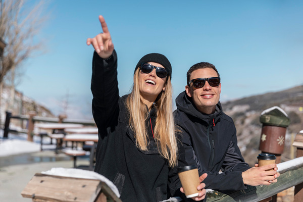 Pareja de turistas apoyados en una valla con vasos de café en la mano, sonriendo en un día soleado en un paisaje nevado.