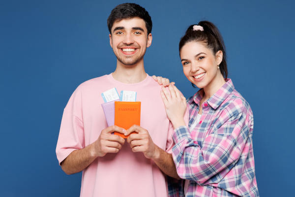 Hombre y mujer sonrientes sosteniendo pasaportes y boletos de avión sobre un fondo liso azul.