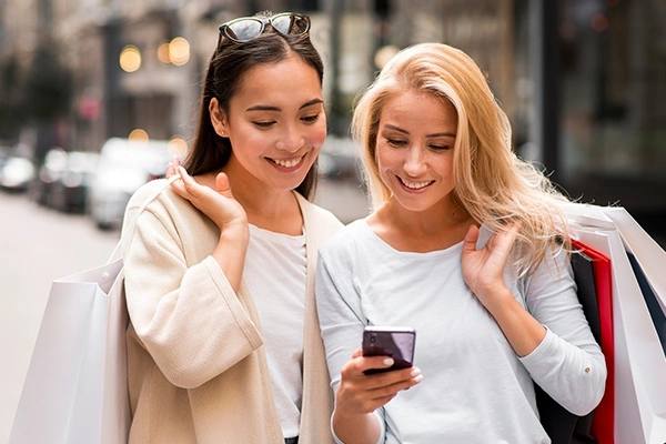 Duas amigas olhando para a tela do celular enquanto fazem compras