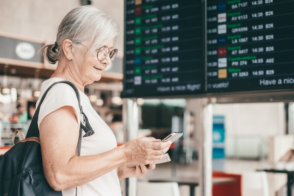Mujer mayor en el aeropuerto usando un smartphone con tablero de entradas y salidas de fondo.