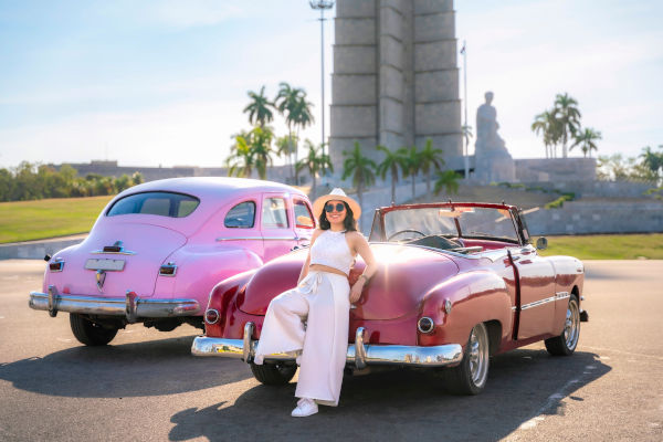 Joven sonriente con gafas de sol y conjunto blanco sentada en un convertible rojo vintage, con el Monumento a José Martí al fondo en La Habana, Cuba.