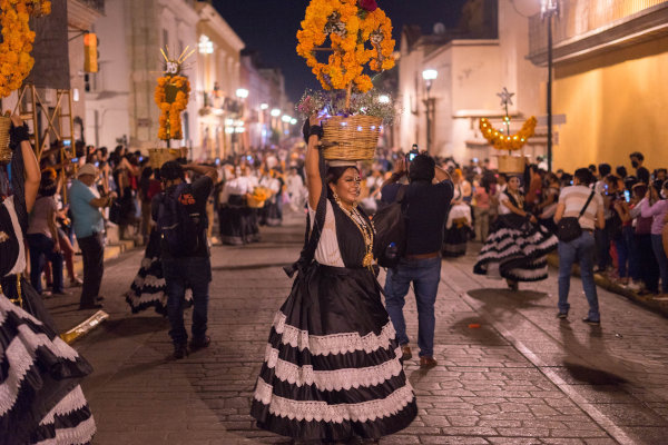 Mujer con vestido blanco y negro y media cara maquillada como catrina danzando con ofrendas en un desfile nocturno del Día de Muertos en Oaxaca.