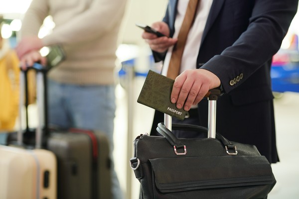Empresario con pasaporte en la mano sobre su maleta de viaje mientras espera en el aeropuerto revisando el teléfono móvil con la otra mano.