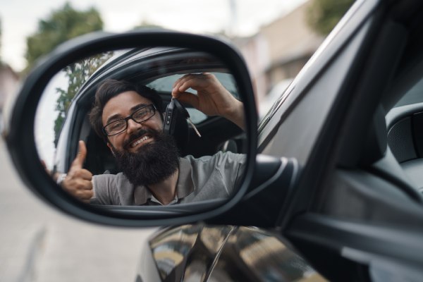 imagem de homem feliz, seguro uma chave de carro, dentro de um veículo e olhando pelo retrovisor para falar sobre dirigir nos eua