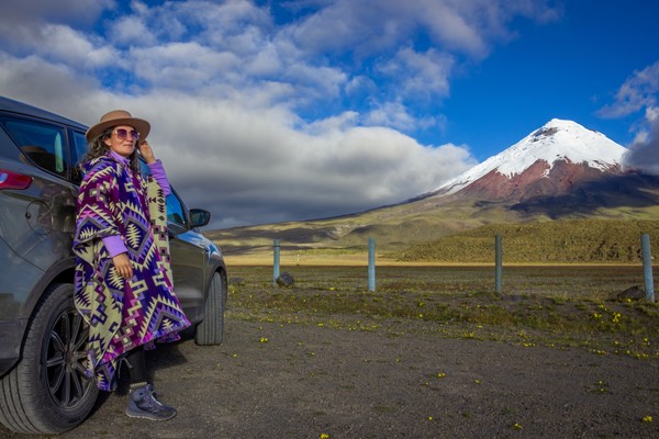 Turista con poncho y sombrero posando junto a su vehículo todoterreno durante un viaje por carretera en el Parque Nacional de Cotopaxi, Ecuador, con el majestuoso volcán Cotopaxi al fondo.