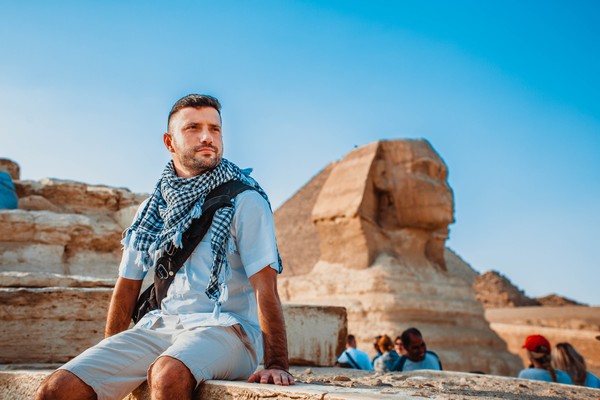 Un joven turista posa sentado para una foto con la Esfinge de Giza de fondo en los alrededores de El Cairo, Egipto