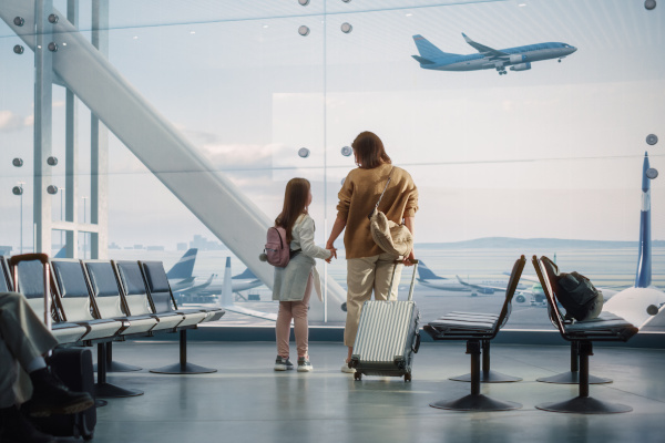 Madre e hija de la mano con una maleta y bolso de mano esperando su vuelo en la sala de espera de un aeropuerto con vista a los ventanales y aviones detenidos de fondo.
