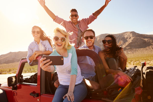 Grupo de personas sonrientes en un viaje por carretera tomándose una selfie sobre una camioneta descapotable en un día soleado.
