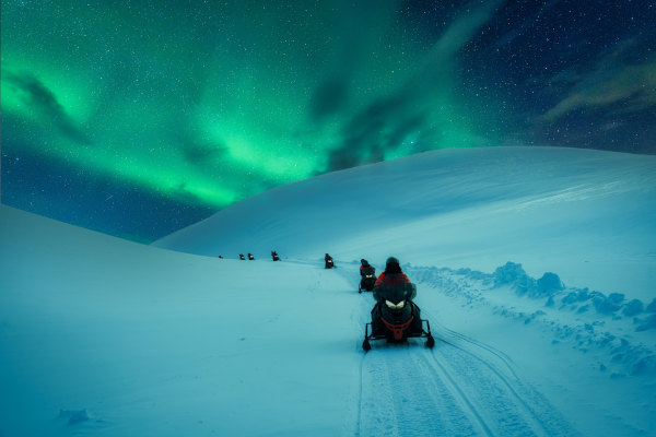 Grupo de viajeros en motos de nieve recorriendo un glaciar bajo una aurora boreal en Islandia.
