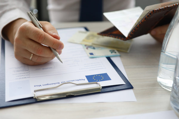 Mano de hombre con bolígrafo rellenando formulario físico de solicitud de visa Schengen en inglés con bandera de la Unión Europea impresa en el papel.