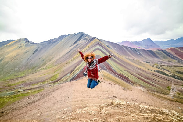 Joven sonriente de cabello rojo saltando con los brazos abiertos frente a la Montaña Arcoiris Vinicunca, Perú.