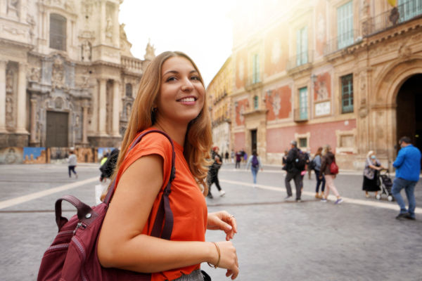 Joven viajera sonriendo con mochila al hombro mientras recorre una plaza histórica en España.