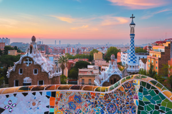Vista panorámica del Parque Güell en Barcelona al amanecer, con sus coloridos mosaicos en primer plano y la ciudad al fondo.