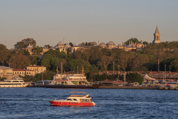 Vista panorámica del Palacio de Topkapi rodeado de exuberante vegetación y edificios históricos, con barco navegando por el Bósforo en primer plano.