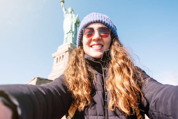 Mujer tomándose una selfie sonriendo con la Estatua de la Libertad en el fondo