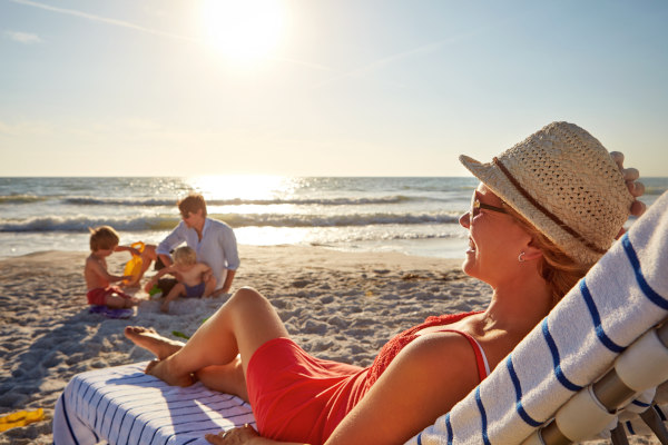 Mujer echada en una reposera en la playa en vacaciones o fin de semana con esposo e hijos jugando de fondo.