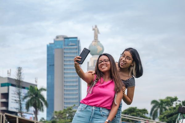 Chicas turistas en la ciudad de El Salvador con edificios y un monumento detrás de ellas en un día nublado.