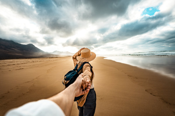 Chica viajera con sombrero y mochila llevando de la mano a su pareja masculina por una playa de arena dorada en un día nublado.
