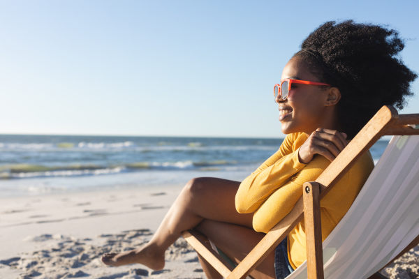 Mujer feliz con gafas de sol sentada en una tumbona blanca sonriendo en una playa en un día soleado.