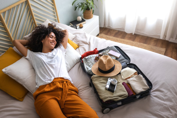 Mujer sonriente echada en la cama con una maleta abierta al lado y múltiples elementos de viaje: sombrero, cámara fotográfica, lentes de sol, anotador y ropa.