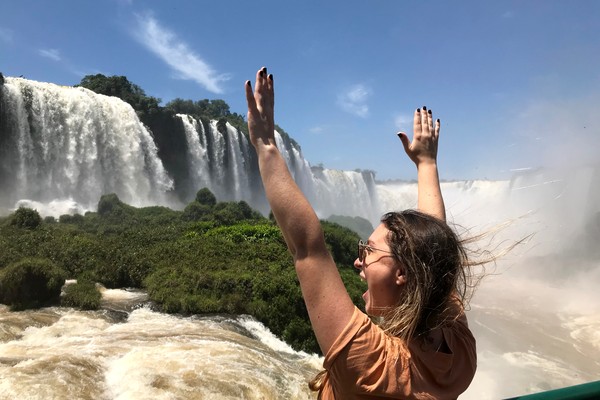 Turista feliz con los brazos elevados en una pasarela frente a las Cataratas del Iguazú del lado brasileño con vegetación de fondo.