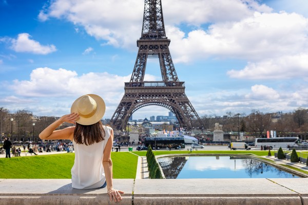 Mujer turista con sombrero sentada en una pared en las cercanías de la torre Eiffel contemplando el monumento con gente alrededor.