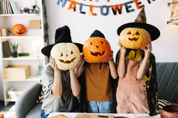 Familia con sombreros de bruja tapándose las caras con calabazas talladas en el interior de una casa decorada para Halloween