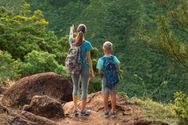 Mujer y niño con mochilas observando el paisaje montañoso en un sendero rodeado de vegetación en Hawaii.