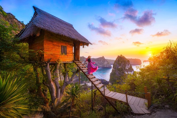 Mujer viajera viendo el amanecer desde la escalera de una cabaña sobre un árbol en los alrededores de la playa Atuh, en la isla de Nusa Penida, Bali, Indonesia.