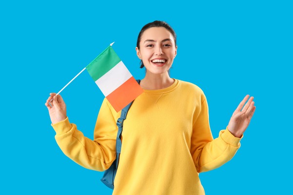 Mujer feliz con suéter amarillo alzando bandera de Irlanda con fondo celeste liso.