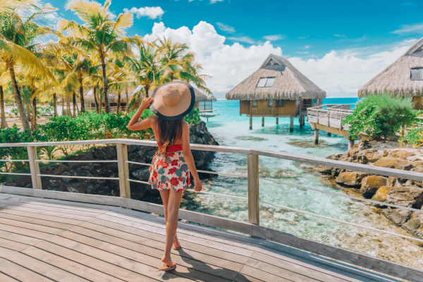 Mujer turista con sombrero y vestido corto mirando bungalows cercanos sobre el mar desde una barandilla en Bora Bora, Polinesia Francesa.