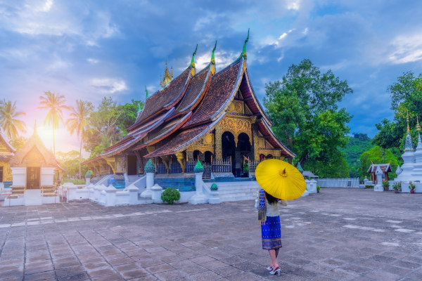 Mujer vestida con traje tradicional de Laos camina por el templo Wat Xieng Thong en Luang Prabang, rodeada de arquitectura budista dorada.